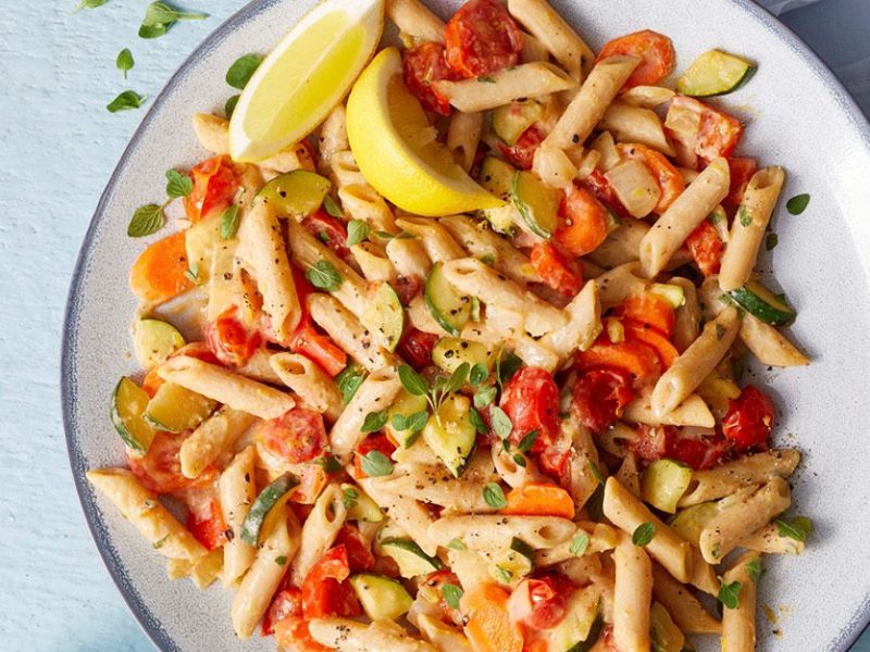 Lemon Hummus Pasta: Penne pasta tossed with a creamy hummus-based dressing, tossed with chopped red bell pepper. Served on a white plate, with lemon wedges on the side.
