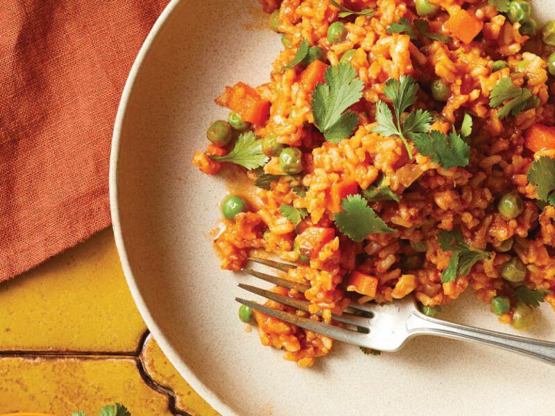 Mexican-Style Brown Rice on a while plate with a metal fork sitting on a yellow tile tabletop