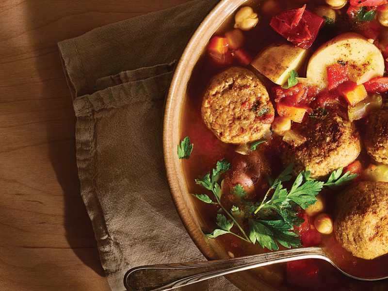 A rustic brown serving bowl of Vegan Kefta Tagine (Moroccan Meatballs) on a brown cloth napkin, with a spoon in the bowl