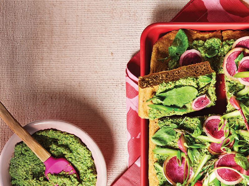 Spring Vegetable Focaccia in a red baking dish, topped witih colorful watermelon radishes and a herbaceous pea spread, next to a small bowl of the green spread