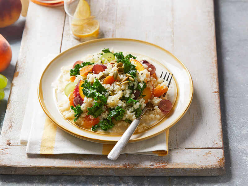 A plate of Couscous and Kale Breakfast Salad on a wooden table with fresh grapes and a peach