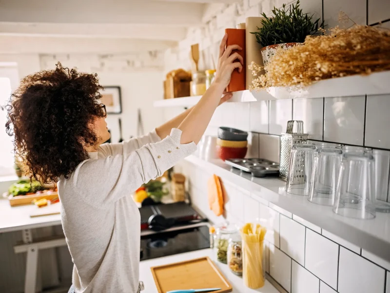 Woman in kitchen reaching for cookbook off shelf