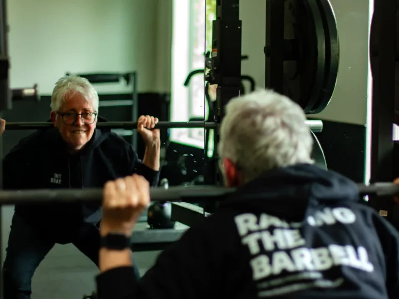 Vegan nun Sister Pat Farrell dead lifts weights in a gym, smiling at herself in the mirror