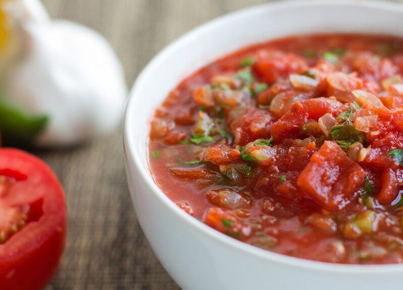 A bowl of tomato sauce with finely chopped fresh herbs to garnish, on a counter next to fresh tomatoes and a bulb of garlic