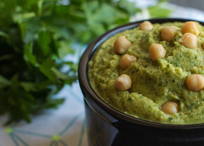 Herbed Hummus in a small black serving bowl, garnished with whole chickpeas and with a bunch of flat-leaf parsley in the background