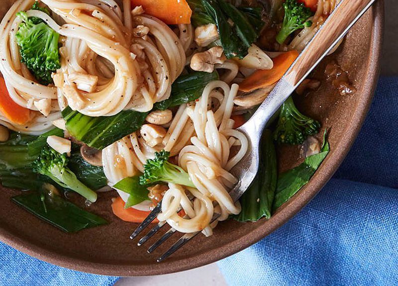 A wide-rimmed brown bowl of Chinese Noodles in Ginger Garlic Sauce next to a blue cloth napkin
