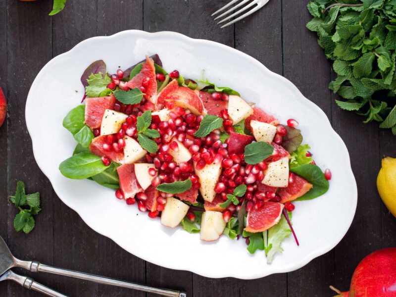 Apple, Grapefruit, Pomegranate Salad on a large, white oval serving platter next to a bunch of fresh mint, a lemon, and fresh whole pomegranate