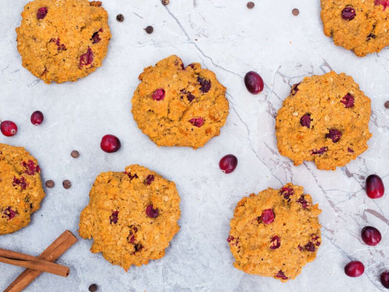 Pumpkin Cranberry Cookies spread out on a marble background with cinnamon sticks and fresh cranberries scattered between the cookies