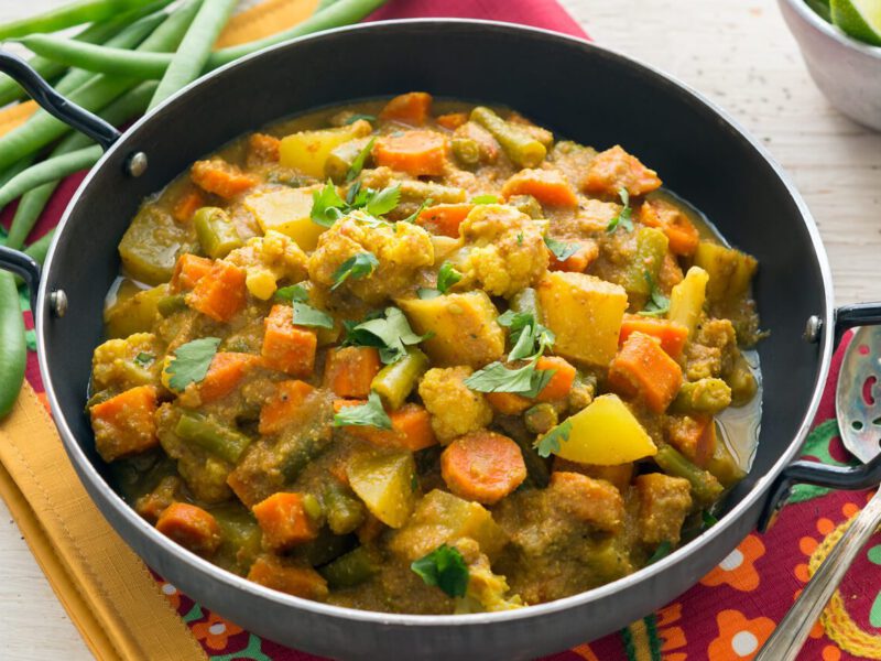 A pan of Mixed Winter Veggie Curry with Poppy Seeds on a colorful placement next to uncooked beans, a bowl of rice, and a cup of lime wedges