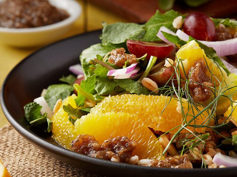 Baby Kale Salad with Farro, Grapes, and Oranges in a black serving bowl with wedges of orange to the sice