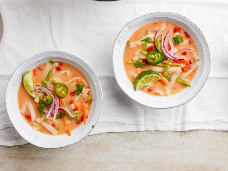 Two colorful bowls of Red Curry Noodle Soup on a white cloth with a small bowl of jalapeno and a bowl of sliced red onion.