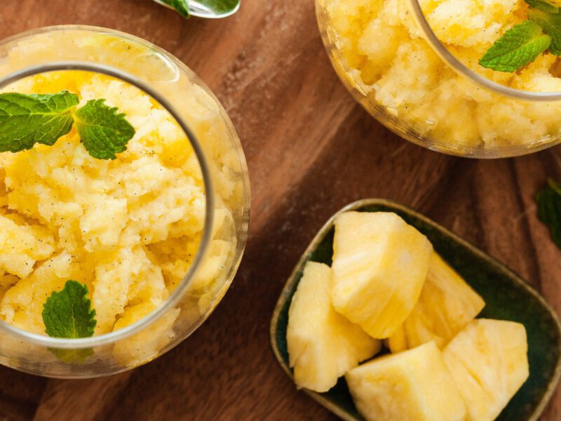 Pineapple-Vanilla Granita in two squat wine glasses on a wooden chopping board with a small bowl of pineapple chunks to the side