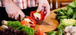 Man's hands holding a knife and chopping a bell pepper on a cutting board, with other vegetables - Plant-based diets have been shown to reduce risks of prostate cancer
